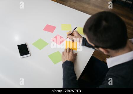 Top view of sticky notes and phone on desk. Office worker holding pen and writing on blank colorful sticky notes on desk with smartphone. Stock Photo