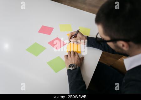 Sticky notes and man's hands on office table. Male hand holding pen writing on empty colorful sticky note with phone on desk. Stock Photo