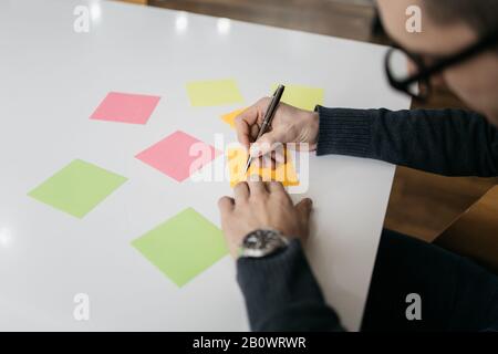 Close-up view of man writing on sticky notes. Sticky notes and man's hands with watch on office white table. Stock Photo