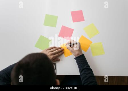 Top view of a white desk with colored sticky notes. Cropped businessman holding pen and writing on sticky notes. Stock Photo