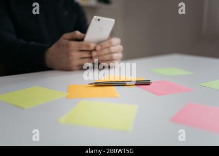 Office desk with sticky notes and pen. Elegant pen on table with sticky notes with blurred men's hands holding phone. Stock Photo