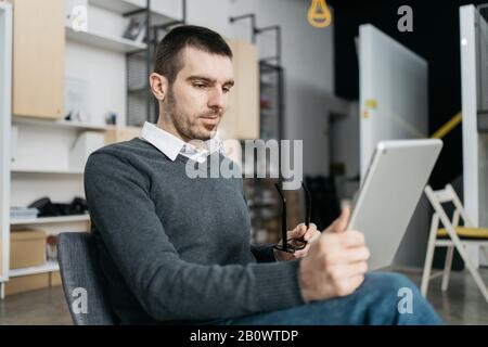 Side view of a man holding eyeglasses and tablet. Office worker looking into a tablet and holding eyeglasses while sitting on chair. Stock Photo