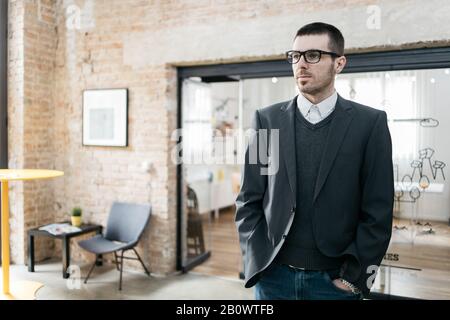 Office guy with grey jacket posing with hands in the pockets. Serious man with elegant jacket standing at office with hands in the jeans. Stock Photo