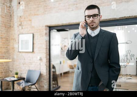 Freelancer posing for photo and looking at camera. Serious man wearing eye glasses and elegant jacket standing at office with hands in the jeans. Stock Photo