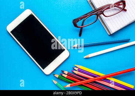 A smartphone on a blue background with colour pencils, notebook and glasses Stock Photo