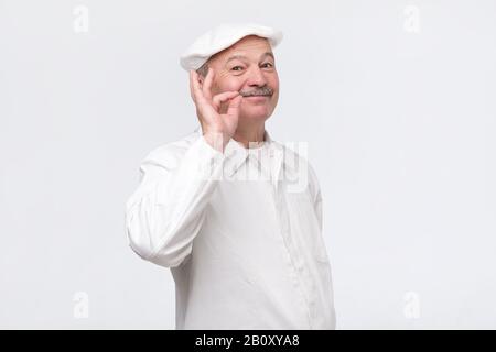 Cooking, food, profession and gesture concept. Smiling senior male chef showing sign ok. Professional male chef in white uniform gesturing excellent. Stock Photo