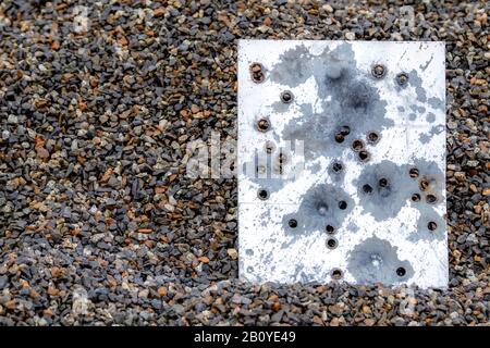 A piece of metal filled with bullet holes. The metal is resting in a pile of dark wet gravel and appears to be the back of a sign. Bullet holes. Stock Photo