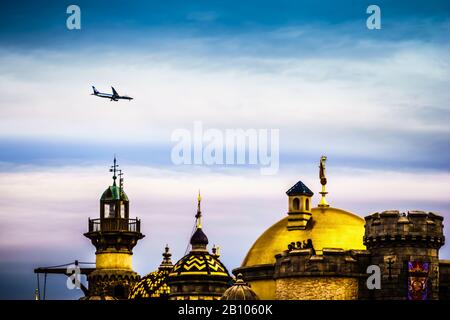 An airplane flying high across a cloudy blue sky over the ornate, golden and striped domes of the Arabian Coast at Tokyo DisneySea. Stock Photo