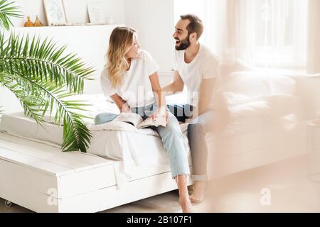 Happy young couple in love relaxing on a couch at the living room, reading magazine, laughing Stock Photo