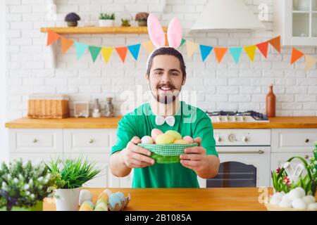 Happy Easter. A funny bearded man holds a basket with eggs and rabbit ears in the decorated room. Stock Photo