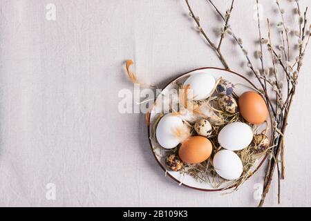 Festive Easter eggs in a clay plate of willow twigs on a table. Light background Happy Easter concept. Flat layout copy space. Stock Photo