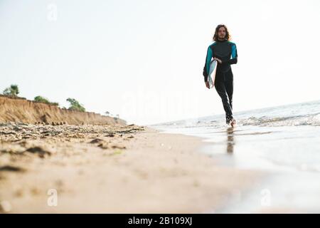 Photo of handsome masculine man in wetsuit holding surfboard while walking at sunny beach Stock Photo