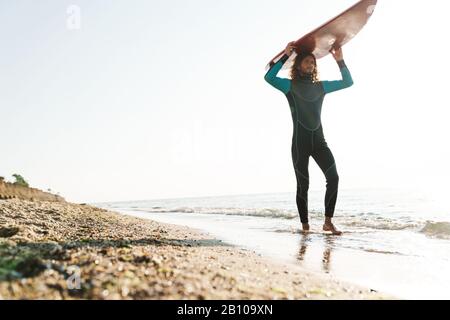 Photo of athletic masculine man in wetsuit holding surfboard on his head while walking at sunny beach Stock Photo