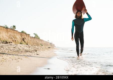 Photo of athletic masculine man in wetsuit holding surfboard on his head while walking at sunny beach Stock Photo