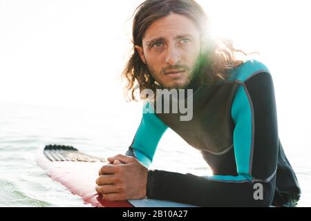 Photo closeup of serious unshaven man in wetsuit using surfboard while working out in sea Stock Photo