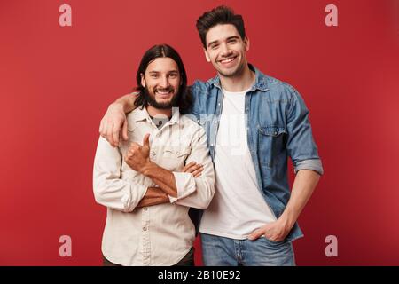 Two cheerful young men wearing casual clothes standing isolated over red background, laughing, hugging Stock Photo