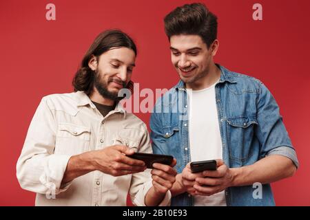 Two cheerful young men wearing casual clothes standing isolated over red background, using mobile phones Stock Photo