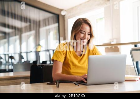 Photo of joyful nice woman using laptop and smiling while sitting at table in open-plan office Stock Photo