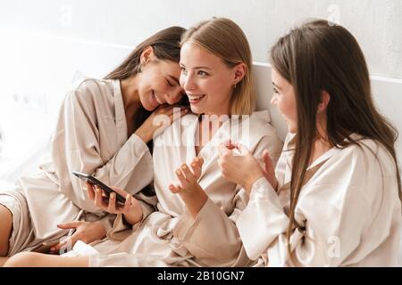 Three cheerful girls friends wearing dressing gowns sitting on bed at home, using mobile phone Stock Photo