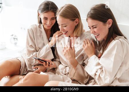 Three cheerful girls friends wearing dressing gowns sitting on bed at home, using mobile phone Stock Photo