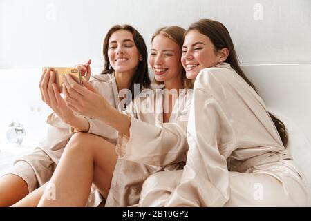Three cheerful girls friends wearing dressing gowns sitting on bed at home, taking a selfie Stock Photo