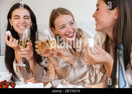 Picture of a cheerful young girls women friends indoors at the kitchen on hen party drinking champagne eat fruits. Stock Photo