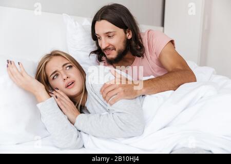 Beautiful young couple laying in bed at home, smiling man waking his confused girlfriend up in the morning Stock Photo