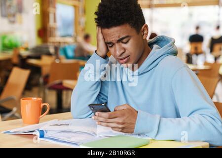 Serious african boy teenager studying while sitting at the hub indoors, using mobile phone Stock Photo