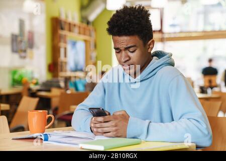 Serious african boy teenager studying while sitting at the hub indoors, using mobile phone Stock Photo