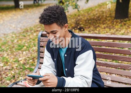 Smiling african boy student sitting on a bench at the park, using mobile phone Stock Photo