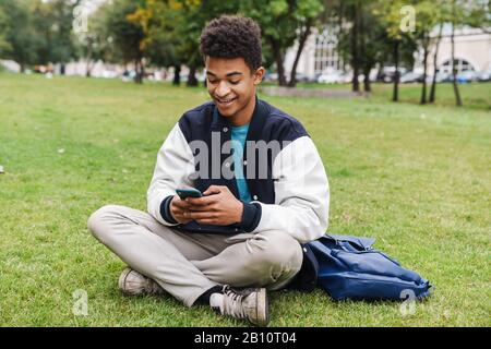 Happy teenager student boy sitting on a lawn at the park, using mobile phone Stock Photo