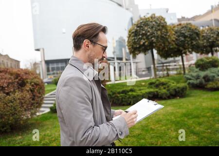 Image of handsome focused businessman in eyeglasses making notes on paper while walking at city street Stock Photo