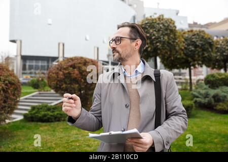 Image of handsome focused businessman in eyeglasses making notes on paper while walking at city street Stock Photo