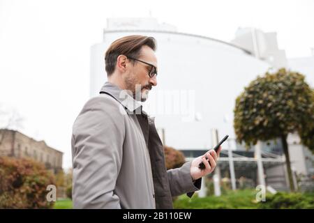 Image of concentrated businessman in eyeglasses using cellphone while walking at city street Stock Photo