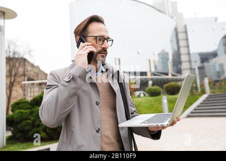 Image of focused bearded businessman in eyeglasses holding laptop and talking on cellphone while standing at city street Stock Photo