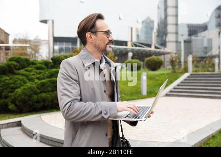 Image of pleased bearded businessman in eyeglasses using laptop while standing at city street Stock Photo