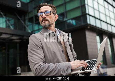 Image of mature bearded businessman in eyeglasses using laptop while standing near office building in urban area Stock Photo