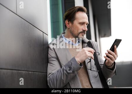 Image of mature confident businessman holding eyeglasses and using cellphone while leaning on wall in urban area Stock Photo
