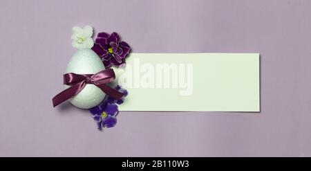 Happy Easter or spring backdrop for banners, greeting cards and web White border with an egg, ribbon and violets on purple background. Stock Photo