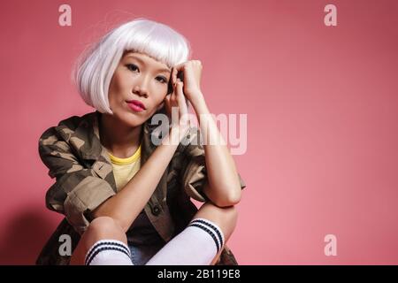 Image of young asian girl wearing white wig sitting and looking at camera isolated over pink background Stock Photo