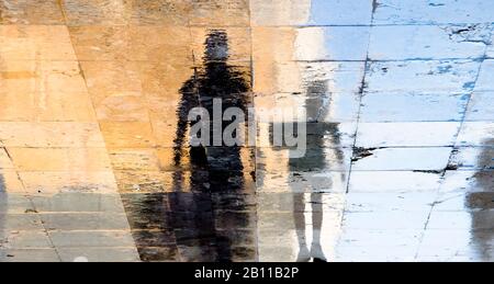 Blurry vintage reflection shadow silhouettes of a man and a boy walking on a wet street on a sunny summer day in old town stone pavement Stock Photo