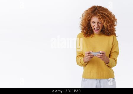 Excited, enthusiastic good-looking redhead curly girl, grimacing having intense game online using mobile device, holding smartphone horizontally Stock Photo
