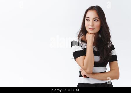 Pensive, attractive and thoughtful young asian brunette woman in striped t-shirt, hold hand under chin, looking away dreamy, smiling contemplating Stock Photo