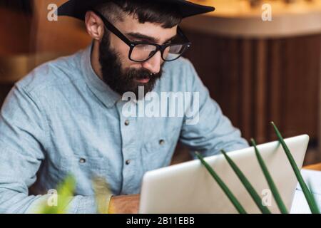 Photo of focused young man wearing eyeglasses using laptop while sitting at table in cafe indoors Stock Photo