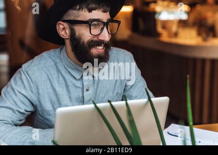 Photo of joyful young man wearing eyeglasses using laptop and smiling while sitting at table in cafe indoors Stock Photo