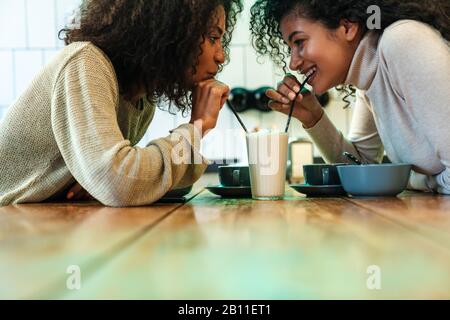 Close up of two happy african girlfriends sitting at the cafe, drinking milkshake from the same glass Stock Photo