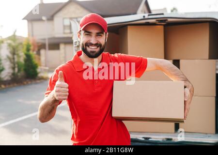Image of smiling young delivery man in red uniform standing with parcel box and showing thumb up near car outdoors Stock Photo