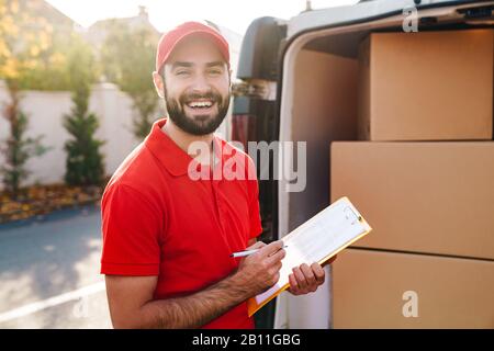 Image of smiling delivery man in red uniform holding clipboard and writing near car with parcel boxes outdoors Stock Photo