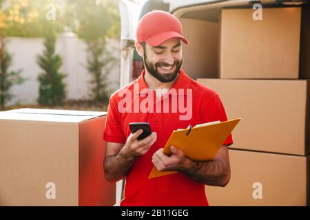 Image of happy delivery man holding clipboard and using cellphone while standing with parcel boxes outdoors Stock Photo