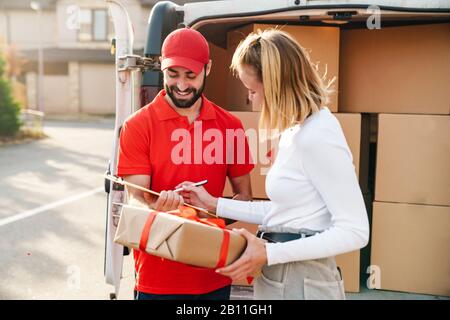 Image of cheerful delivery man in red uniform getting order from caucasian woman near car with parcel boxes outdoors Stock Photo